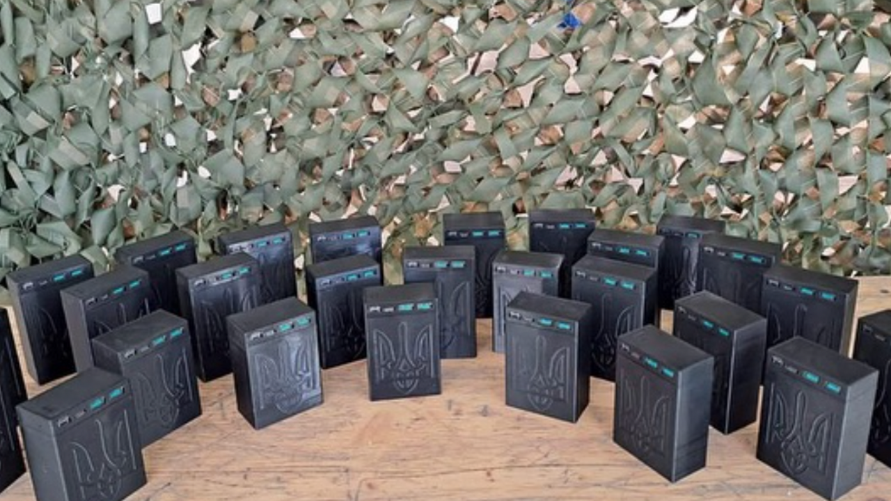 Over two dozen powerbanks displayed in the shape of a crescent on a wooden table. Behind is a textured green backdrop.