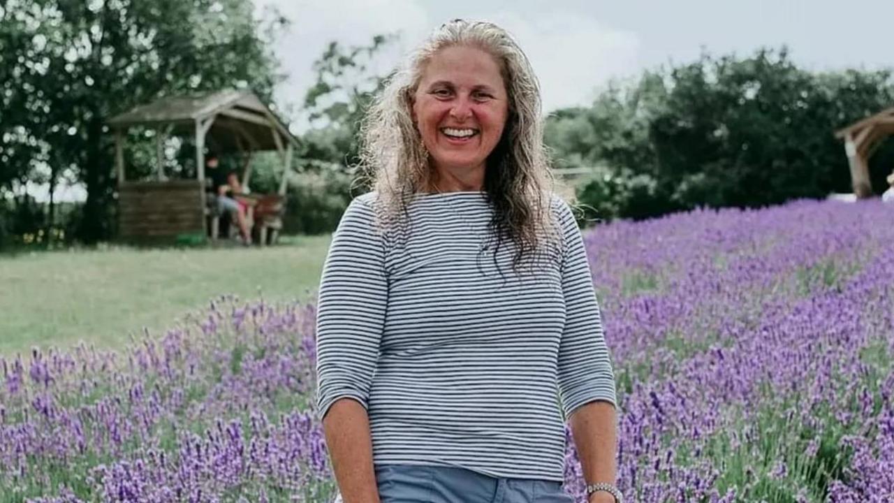 Tina Bessell is surrounded by lavender in bloom. She is smiling and has long pale hair and is wearing a striped top. Behind her, slightly out of focus, is a wooden summer house in which someone is sitting with their back to the camera.