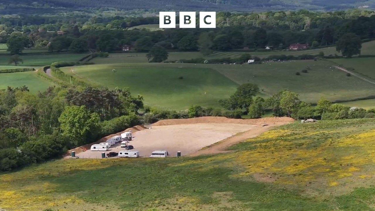 An aerial view of rolling fields and green countryside. In the middle is a freshly dug field, with a hardcore standing and several caravans and trailers parked up.