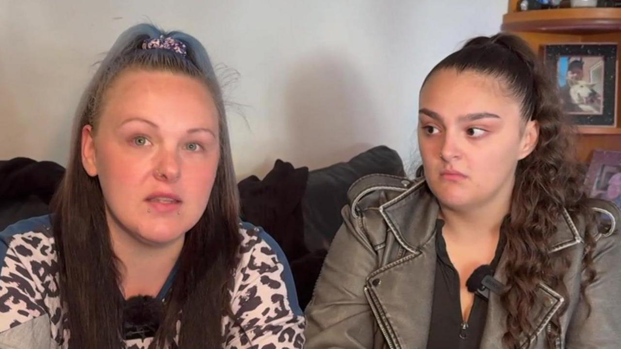 Rebecca and Lucy Barnard are sitting on a sofa together. They both have long dark hair. Lucy is looking at Rebecca as she speaks. A photograph of their brother is on a shelf behind them.