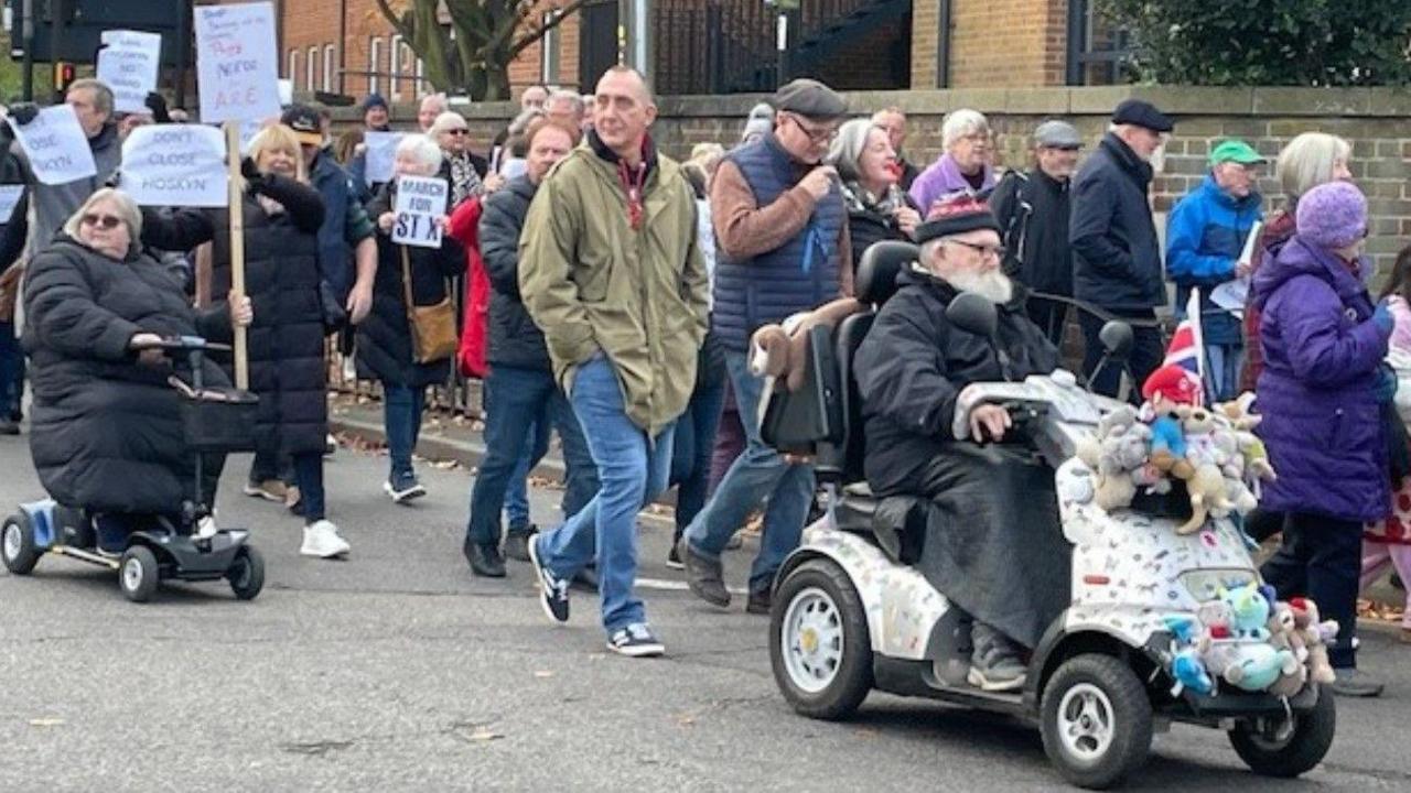 People march, with some carrying a placard or a piece of paper. Some people are walking in the road, along with two people on mobility scooters, and others are on the pavement.