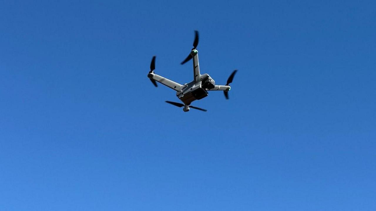A drone in the air. The drone is grey and black with four arms with rotors on the end of them. It is against a purely blue sky, in the air.