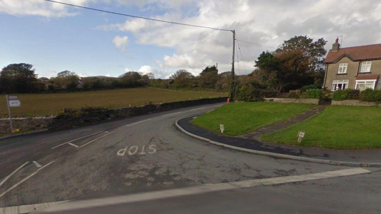 The corner of a road with STOP written on the tarmac, alongside grass verges and nearby house.