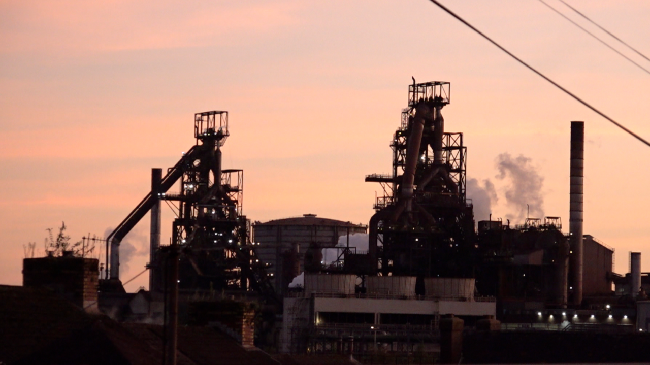 A shot of the steelworks' blast furnaces in Port Talbot. White smoke is coming from the steelworks and a number of other industrial buildings can also be seen. The photo is at dusk and the sky is pink with very few wispy clouds.