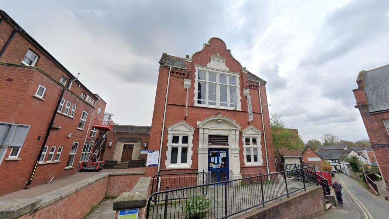 A Google street view image showing a two storey library building in the middle of a town, with a black fence in front of it. A narrow road is on the right with a man walking on the pavement.