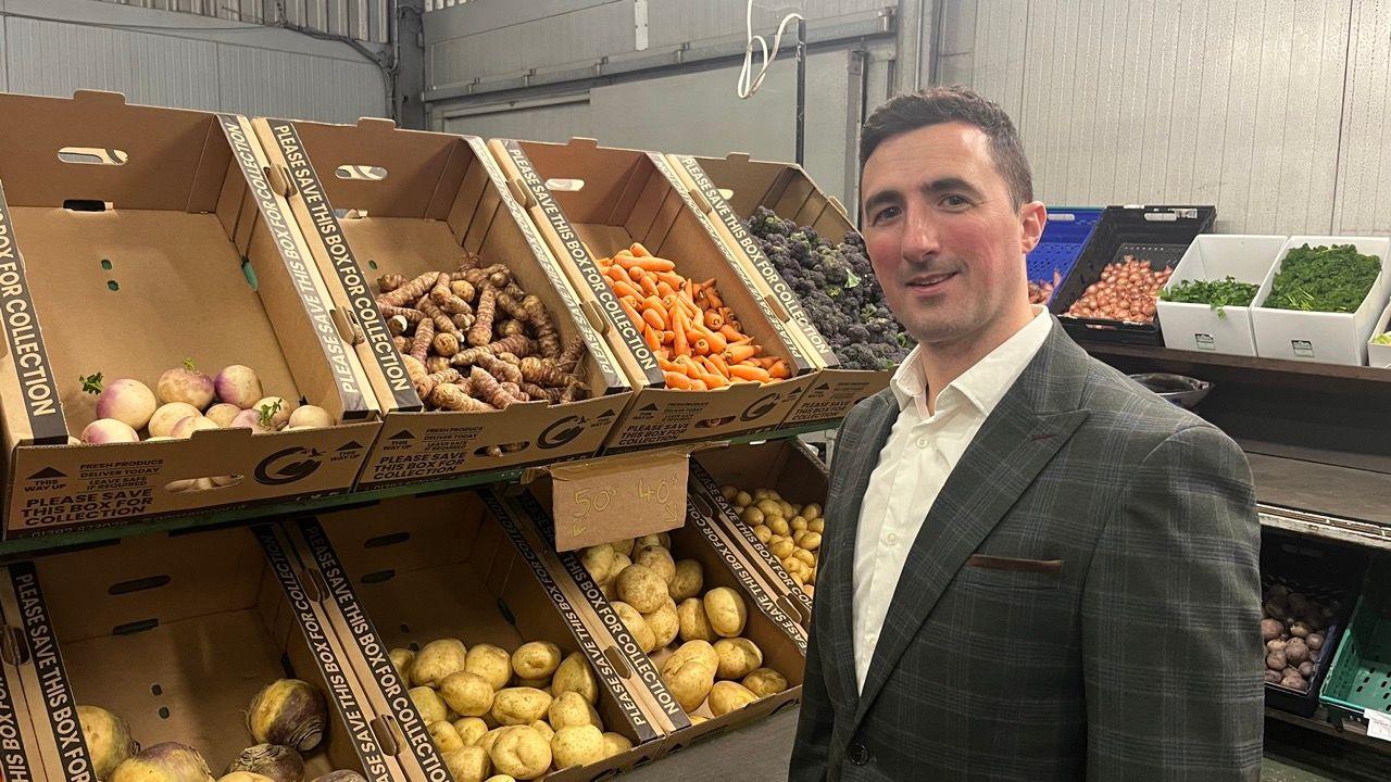 Jamie Walsh wearing a white shirt and a grey check blazer and standing in front of boxes of vegetables in the distribution centre.