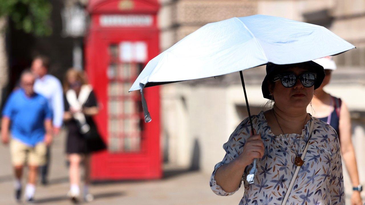 A woman wearing a hat and sunglasses, holding a white umbrella on a sunny day, with three people walking behind her and a red telephone box in distance