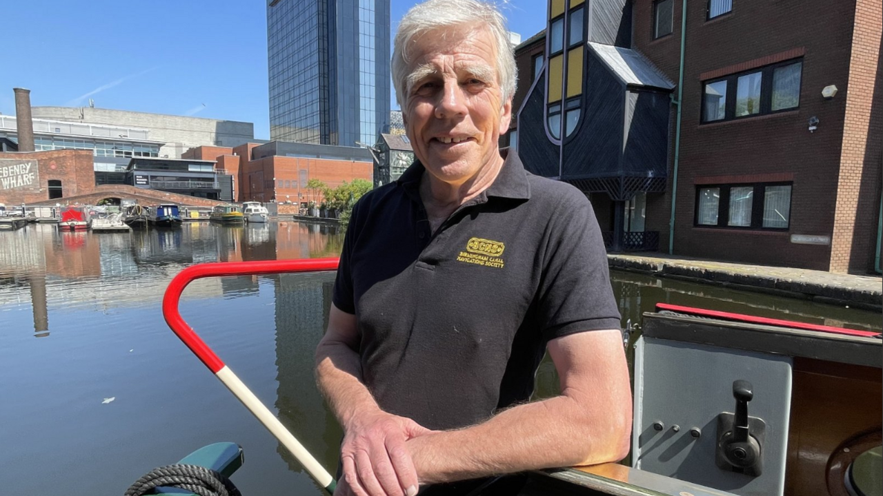 Bob is standing on a canal barge and is wearing a black polo-type shirt. The sun is shining and he's leaning on his elbow and smiling at the camera. He has grey hair.