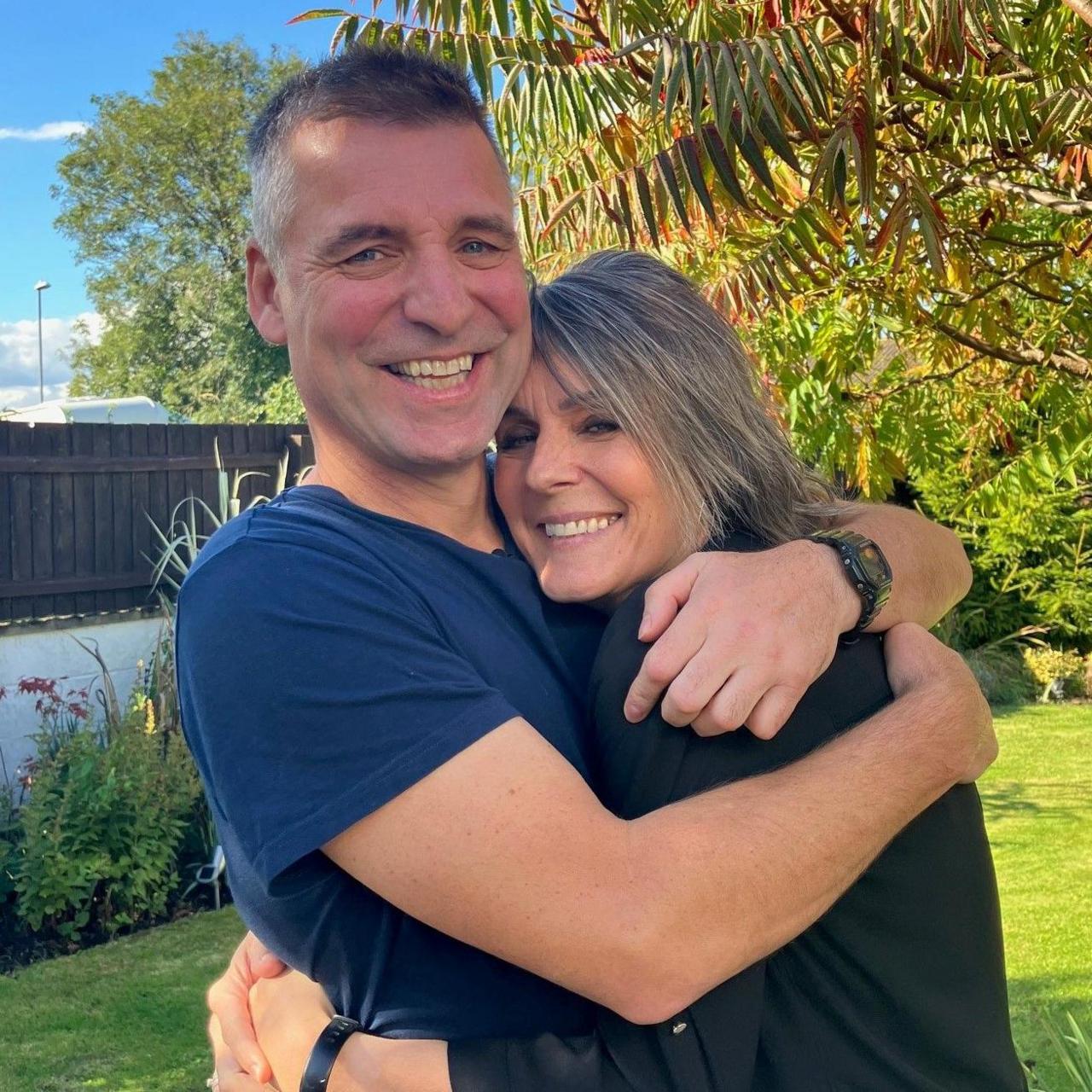 A couple - Jeannie Ambrose and her husband - are hugging in a garden. They both are smiling at the camera.