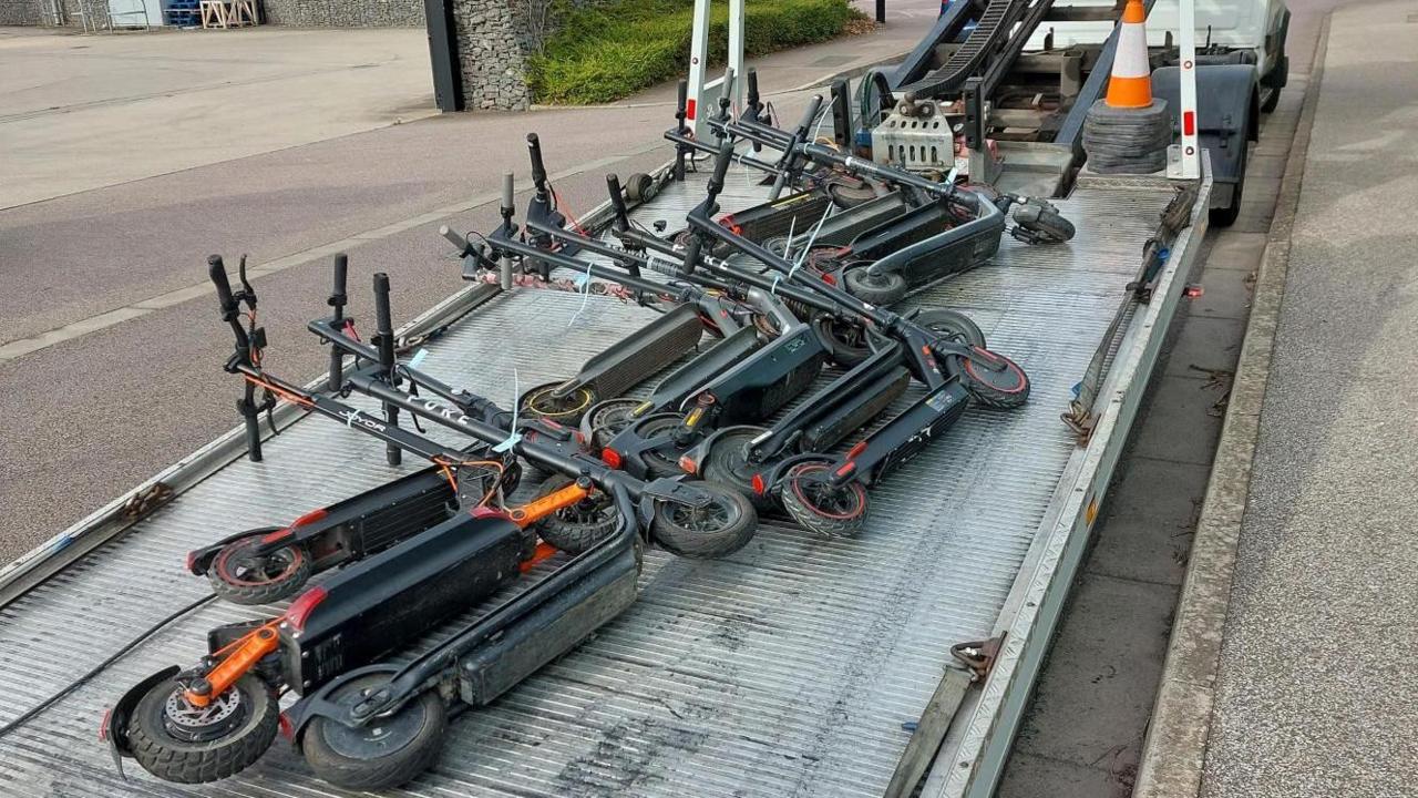 A number of e-scooters are seen on the back of a flat-bed truck which is parked at the side of a road