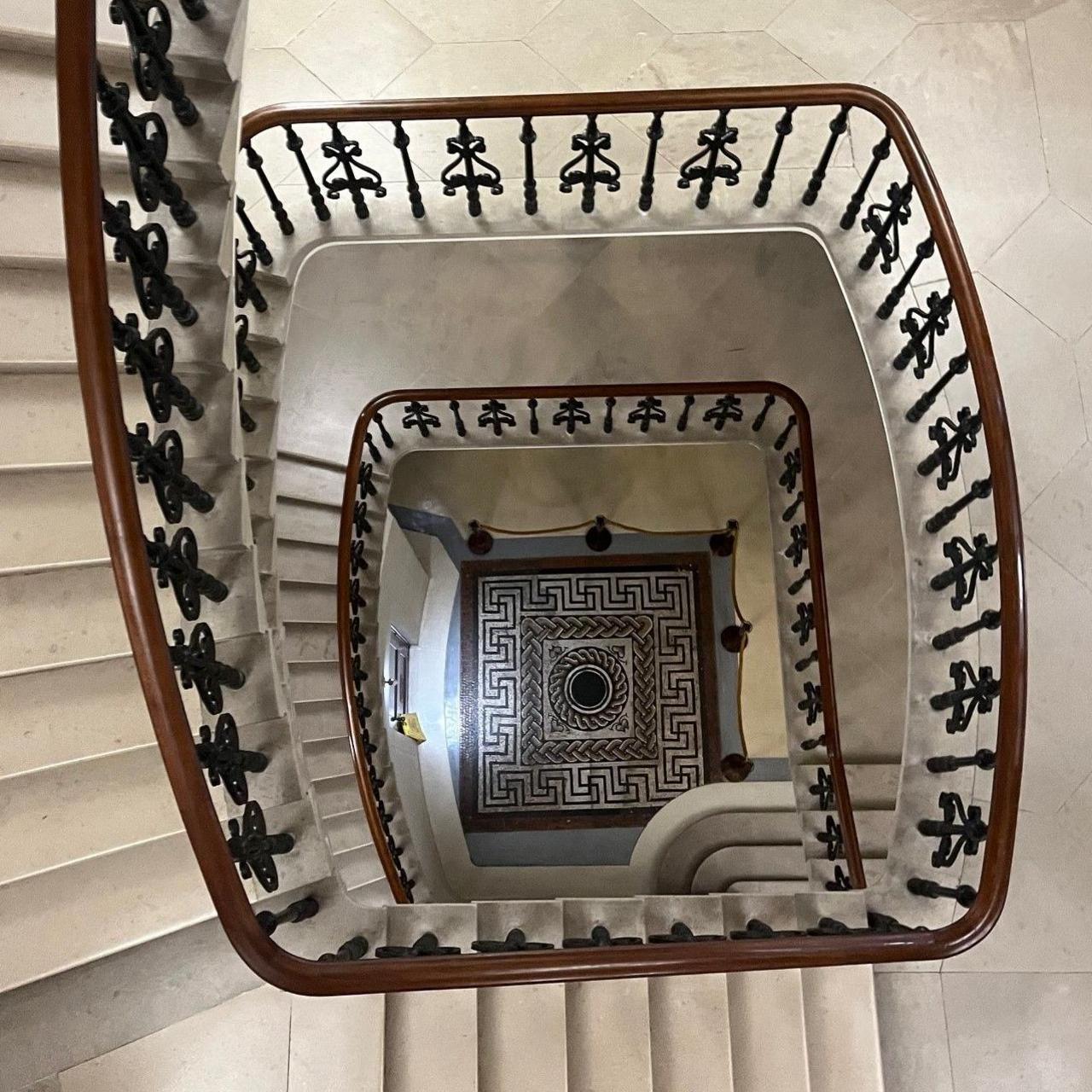 A black, white and grey geometric mosaic at the bottom of a spiralling stone stair case which has a wooden bannister