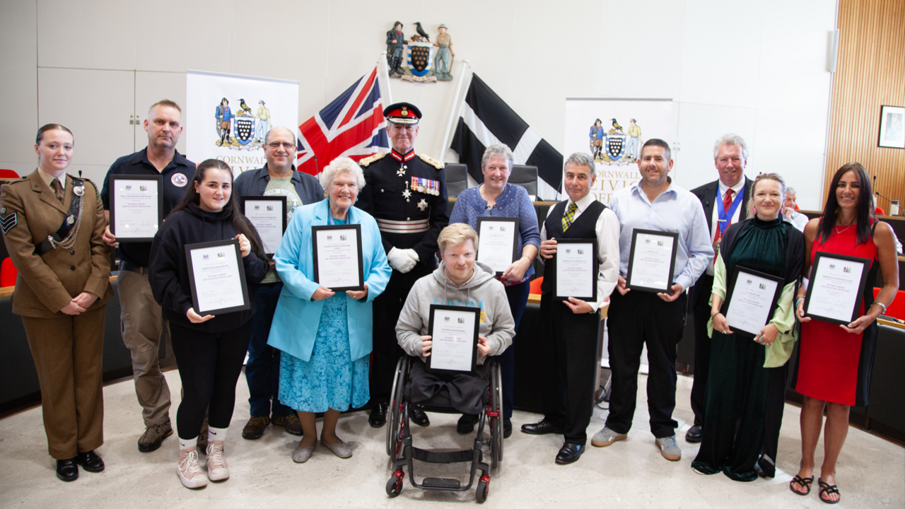 A group of people holding certificates while smiling at the camera. There is a black and white Cornish flag on the right and an England flag on the left behind a man at the centre. 