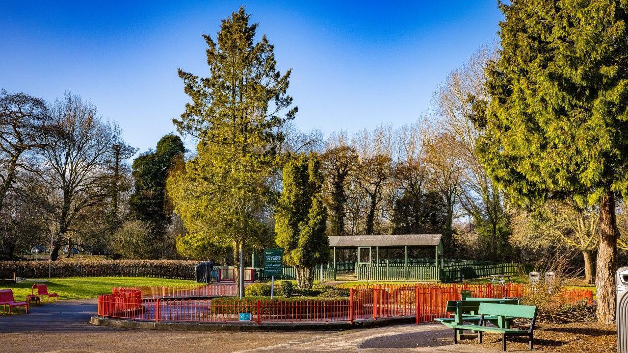 Ground level view of a typical city park, with a number of mature trees among lawn areas and a small pavilion