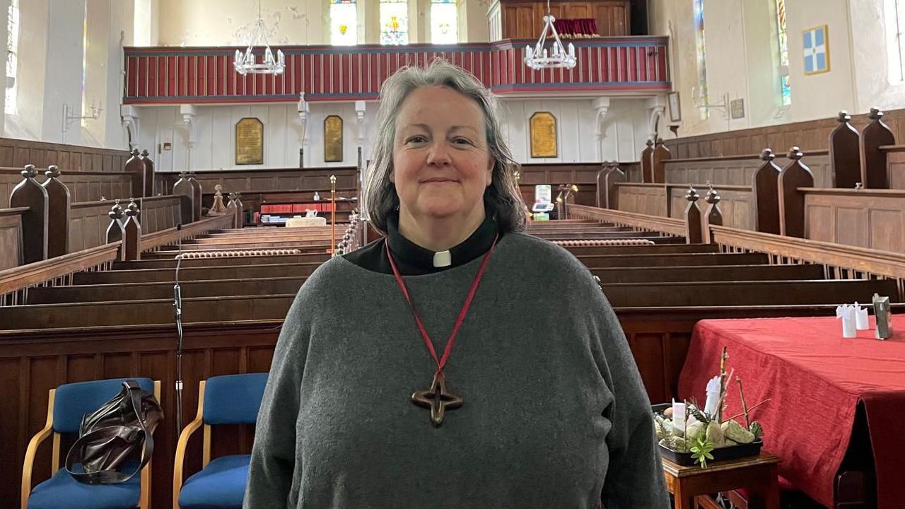 A person with a grey sweater and dog collar with a cross hanging on the chest with a church and pews in the background.