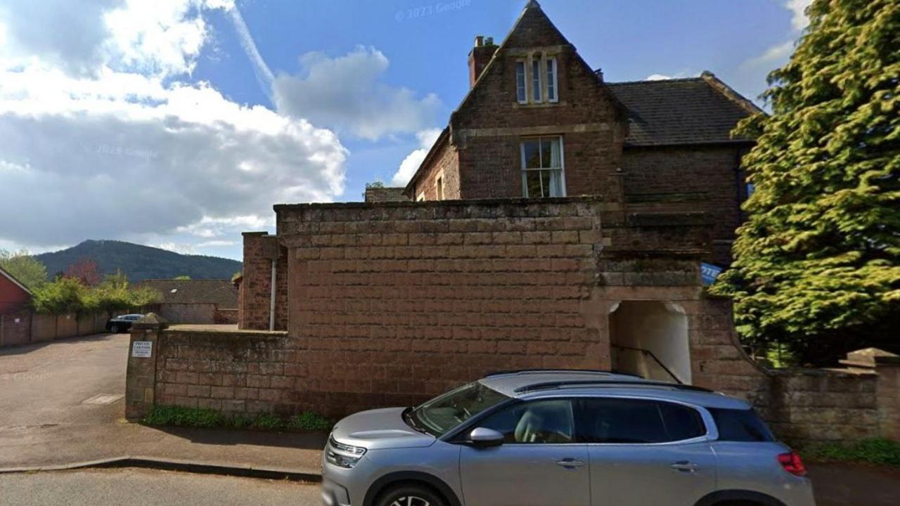 A side view of a Victorian three storey building, partially obscured by a boundary wall. A silver car is parked on the street in front and a carpark lies to its left.