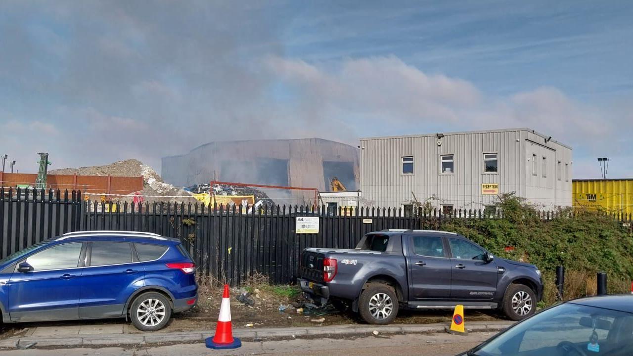 Image of cars parked on the side of the road and smoke coming out of the recycling centre.