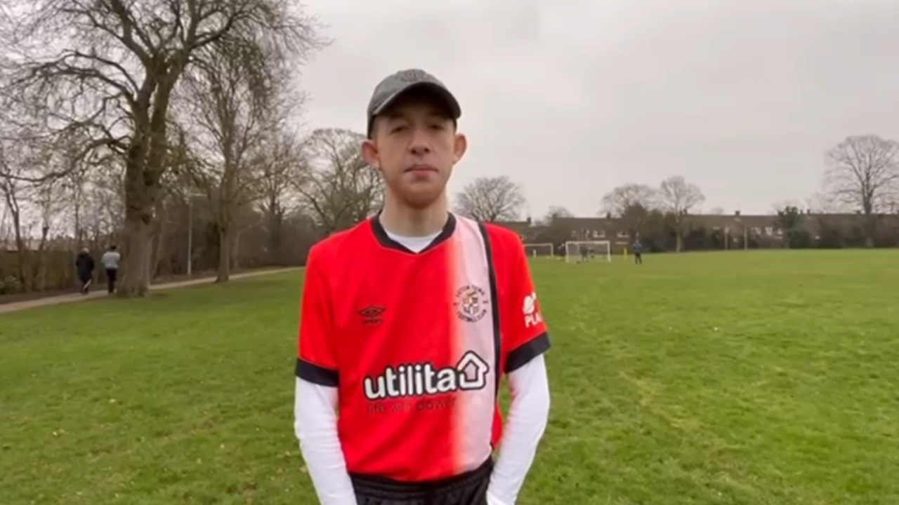 Aidan wearing a red and white Luton Town football shirt looking at the camera. He is standing on a football pitch.