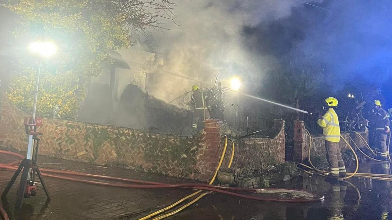 A firefighters aims a water jet hose towards a building which has smoke billowing out of it. The firefighter wears a florescent yellow hat and coat. Other firefighters can be seen in the distance. A portable light has been set up on a driveway.