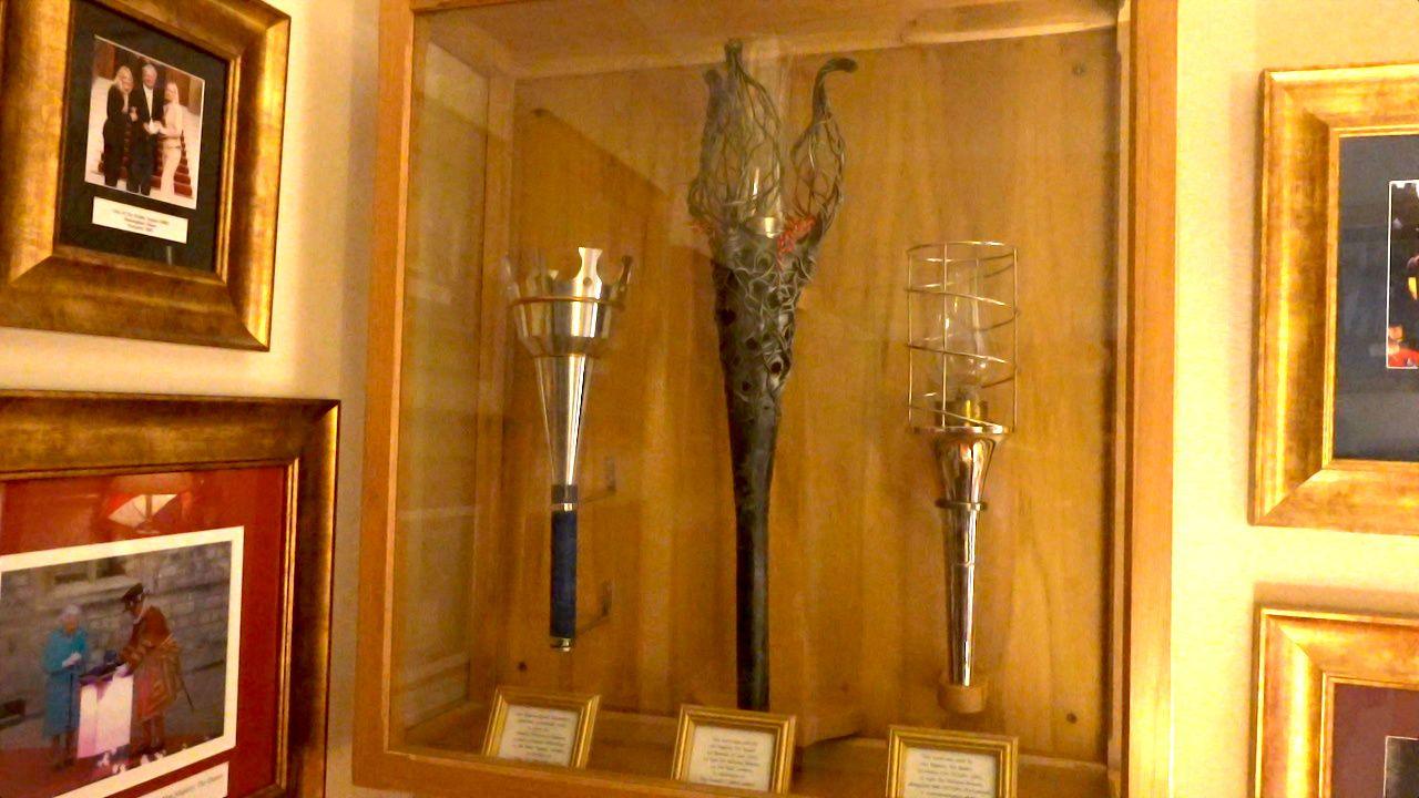Ceremonial torches in an enclosed glass and wooden-framed case are mounted on a wall. Adjacent are gold-framed photos including one of the late Queen Elizabeth II with a Beefeater in his red and gold outfit.