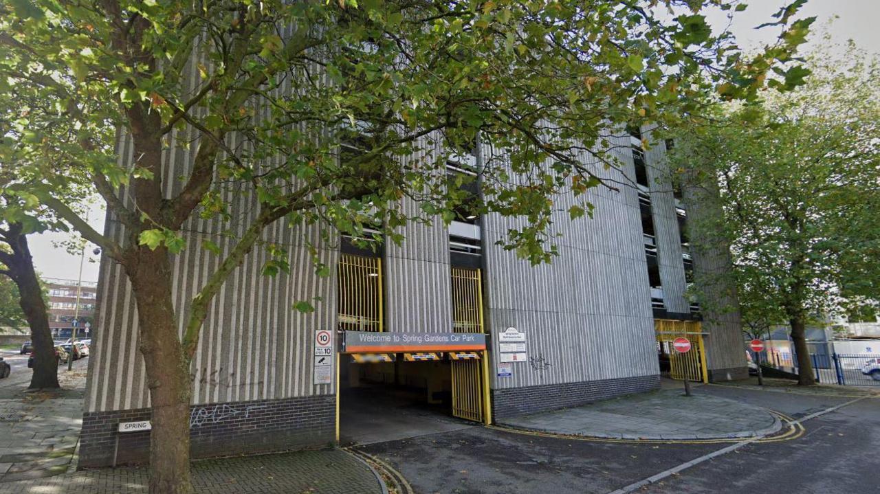 A street view of the entrance to Spring Gardens car park in Swindon. It is a five storey car park. The concrete building has vertical lines going up it and yellow bars near the entrance, which is surrounded by trees.