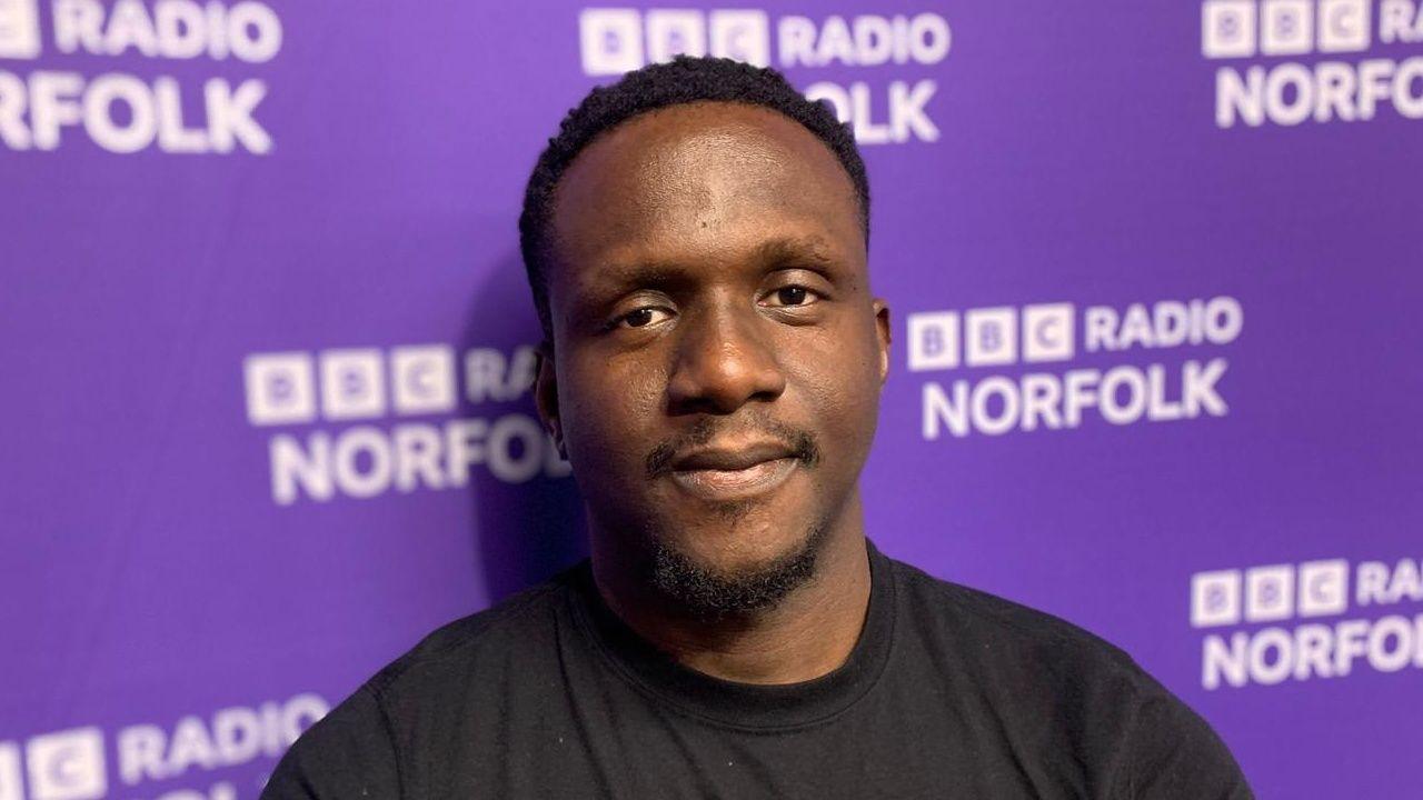 A close-up of Dr Sopuru Mokwe, he's wearing a black T-shirt and standing in from of a purple screen with BBC Radio Norfolk branding.