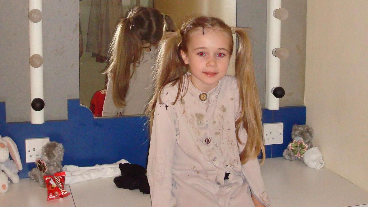 A young girl with pig tails smiles as she sits on a dressing table with her back to a mirror