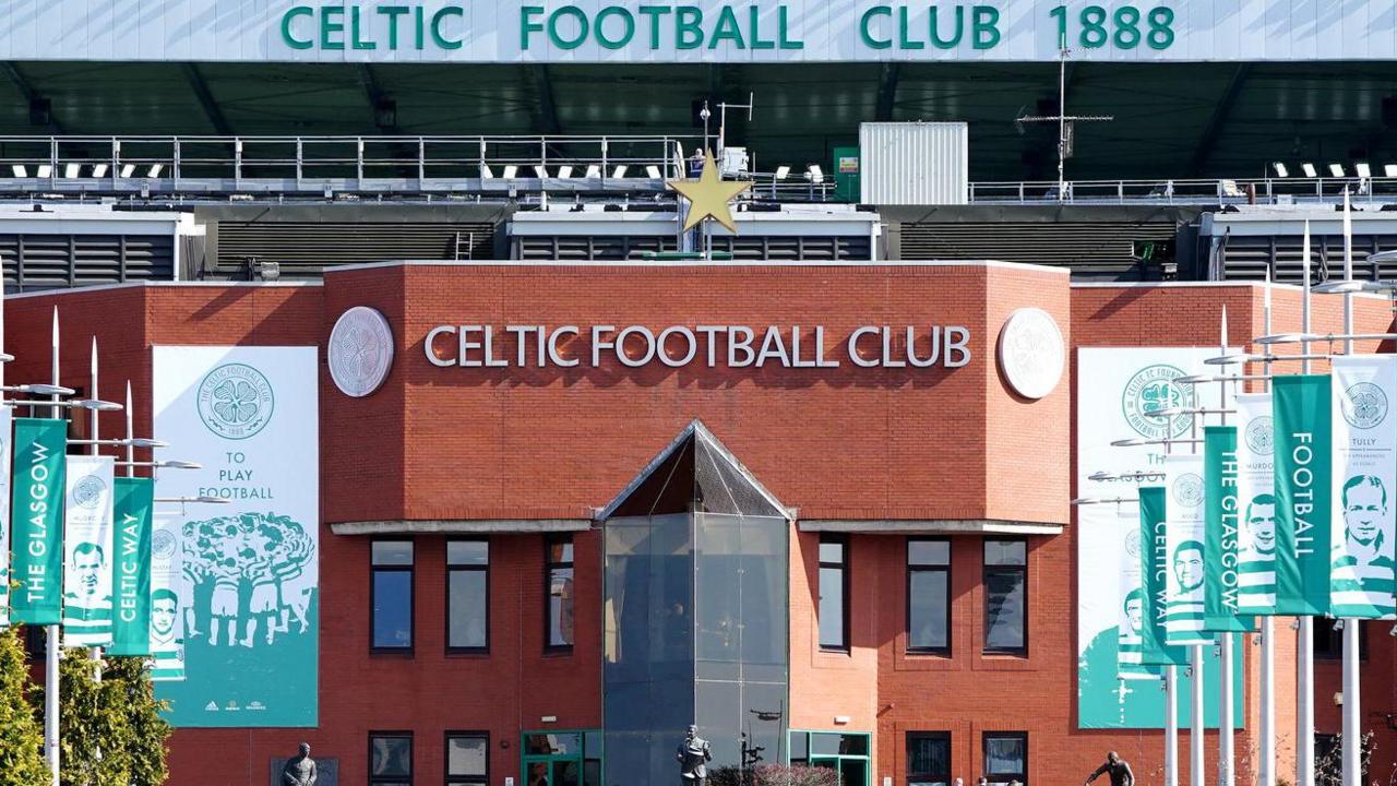 A view of the entrance to Celtic Football Club. It is a redbrick building with a walkway flanked by tall vertical flags showing players and slogans in green and white. A sign at the front top of the building says Celtic Football Club in silver and a gold star sits above the words.