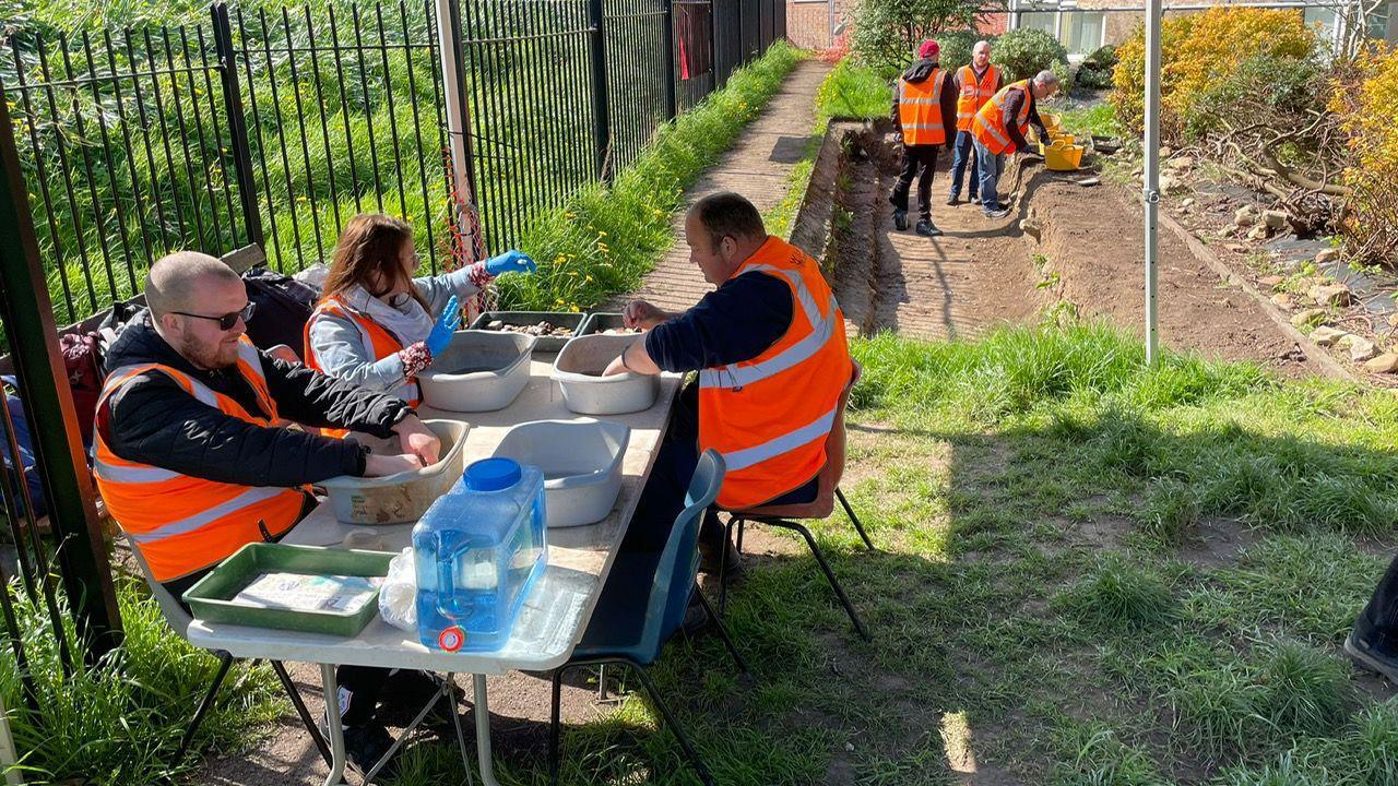 Three people sit at a table outdoors wearing high vis and with their hands in plastic boxes. In the background a section of the ground has been dug out and three people are standing in it.