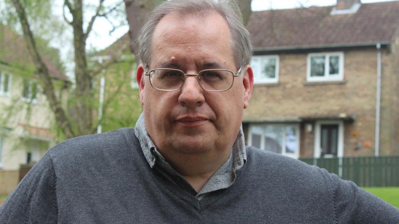 Andrew Rice is looking at the camera. He is wearing a grey jumper on top of a grey shirt and thin-rimmed glasses. There is a house and trees behind him.