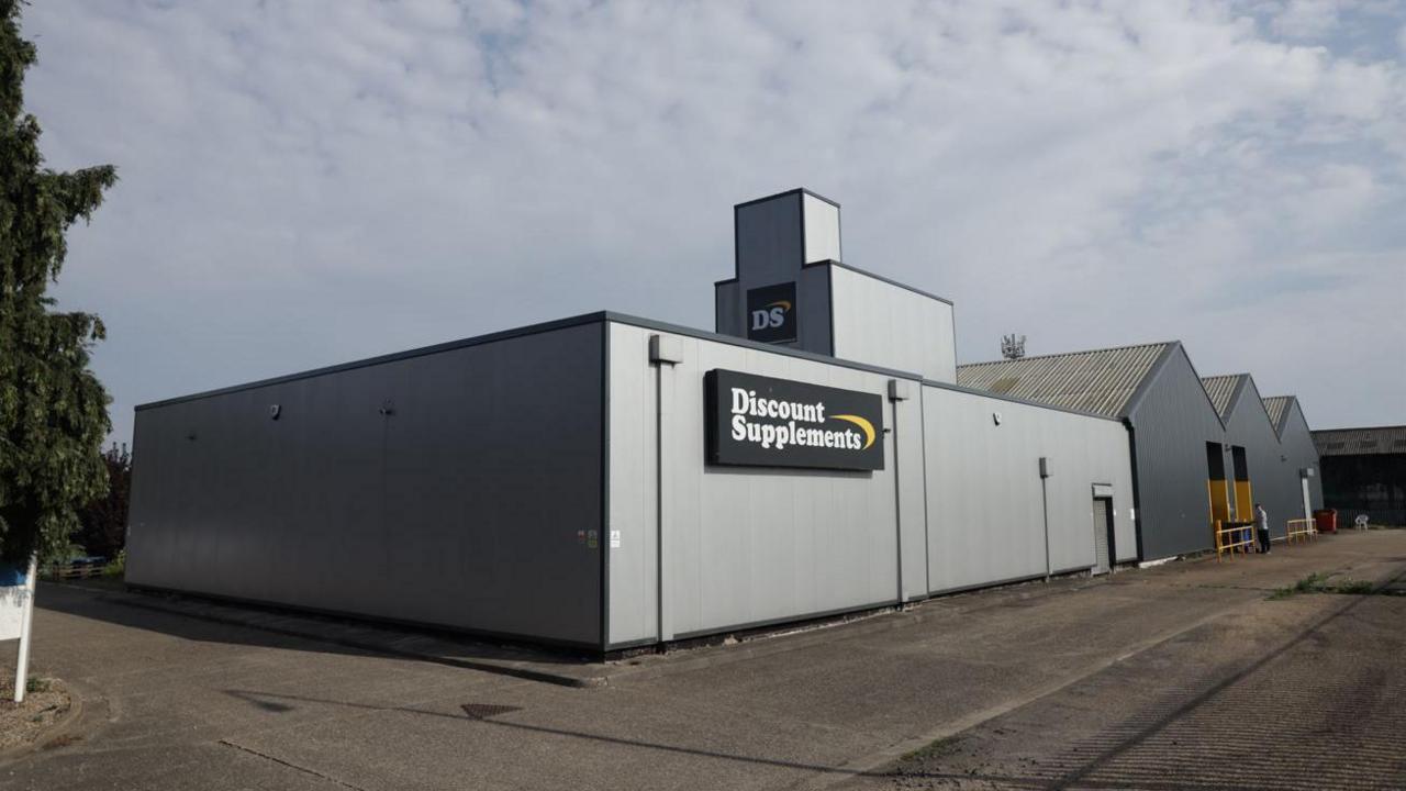 A landscape photo of a rectangular grey warehouse building with three triangular black buildings next to it. The grey building has a black sign on it saying Discount Supplements in white font.