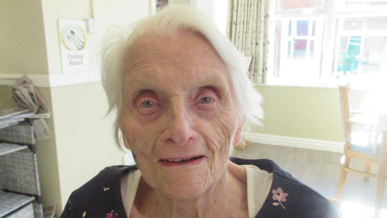 An elderly woman smiling at the camera. She has grey hair and is wearing a white top with a black top over it with pink small flowers. She is inside a living room with chairs and shelves in the background.