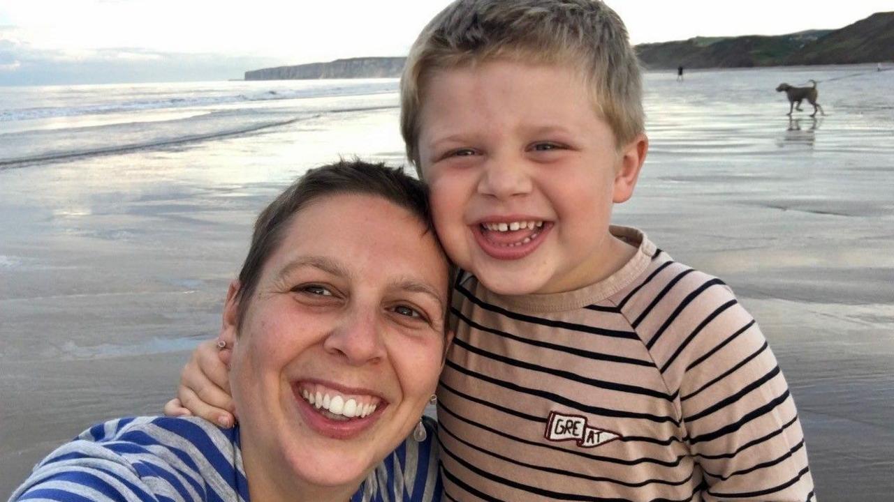 A parent and a child are smiling in a selfie photo at the beach. You can see cliffs and the beach and sea in the background.