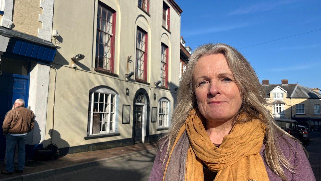 Rachel Kelly wearing an orange scarf and a lilac jacket in front of the Globe Hotel in Torrington