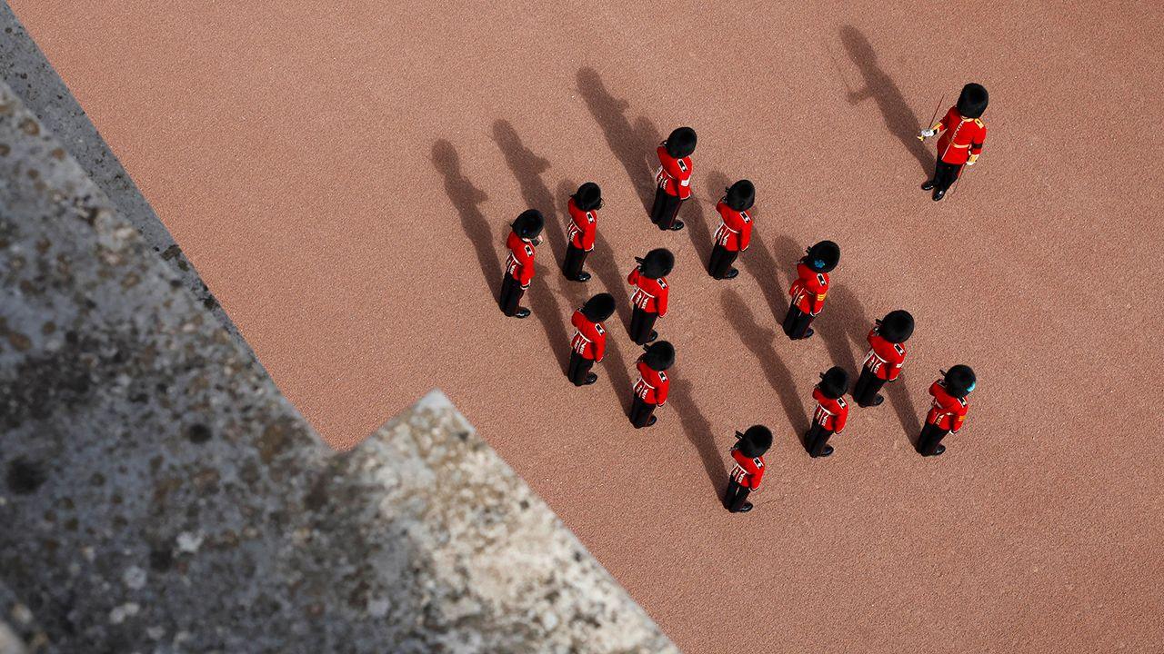 An aerial shot shows Coldstream Guards standing to attention while wearing their distinctive red uniform and black bearskin caps, as a low sun casts long shadows on the tarmac outside Buckingham Palace during the State Funeral of Queen Elizabeth II on September 19, 2022 in London