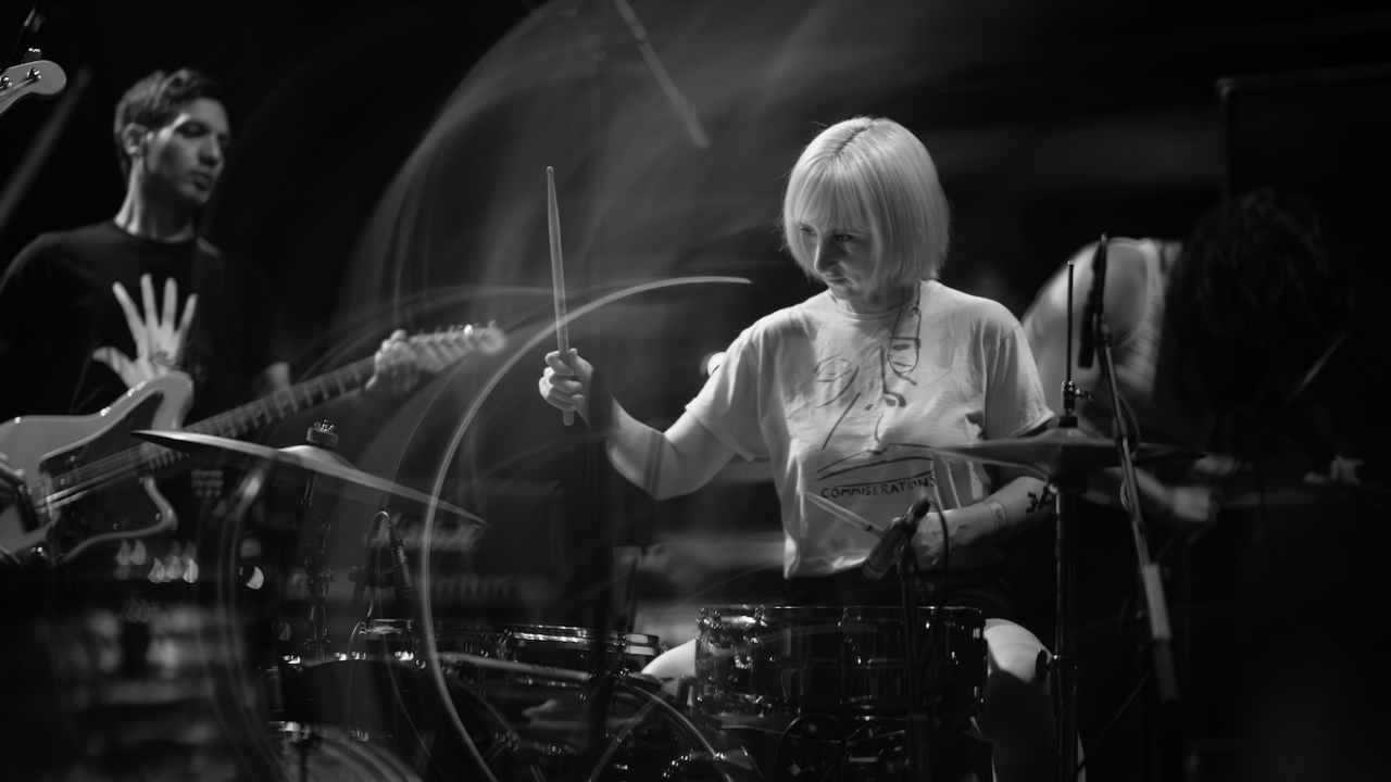 A female member of Water Damage plays drums. She has blonde hair and is wearing a light coloured tee-shirt. A guitarist can be seen in the background. 
