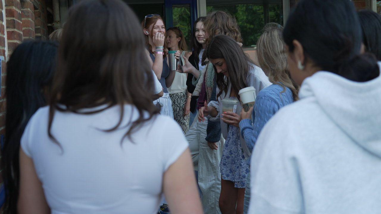A number of young women are standing outside of the colleg waiting to get in for their results. Some of them are looking nervous and quietly waiting, others are smiling and chatting. They are all wearing casual, summery clothes and some are holding takeaway coffee cups.