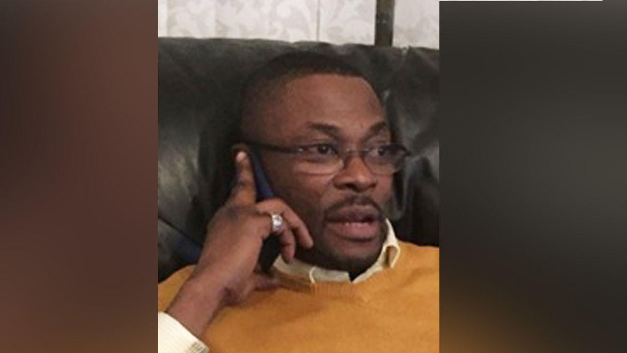 Calvin Olawale Babatunde wearing an orange jumper with a white collared shirt and dark-rimmed glasses. He has a goatee and is holding a phone to his ear.