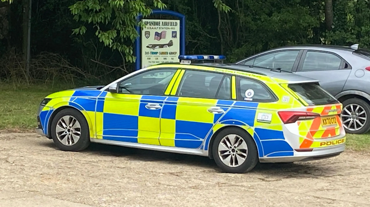 A police car is in the middle of the picture on some brown concrete. It has chequered blue and yellow pattern, and the front of the car is facing towards the left side of the picture. In the background there are lots of trees, and a sign which says Spanhoe Airfield on it. 