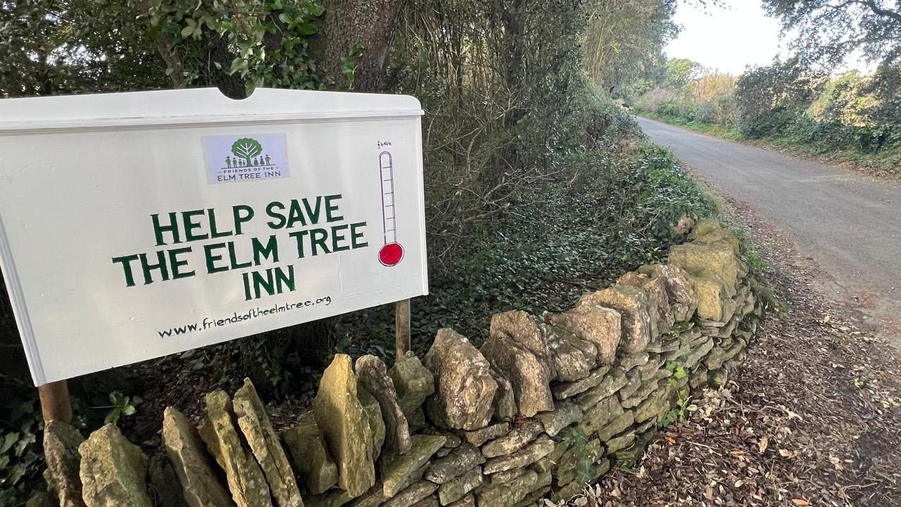 A sign on the a street corner above a dry stone wall which reads 'help save the Elm Tree Inn'.