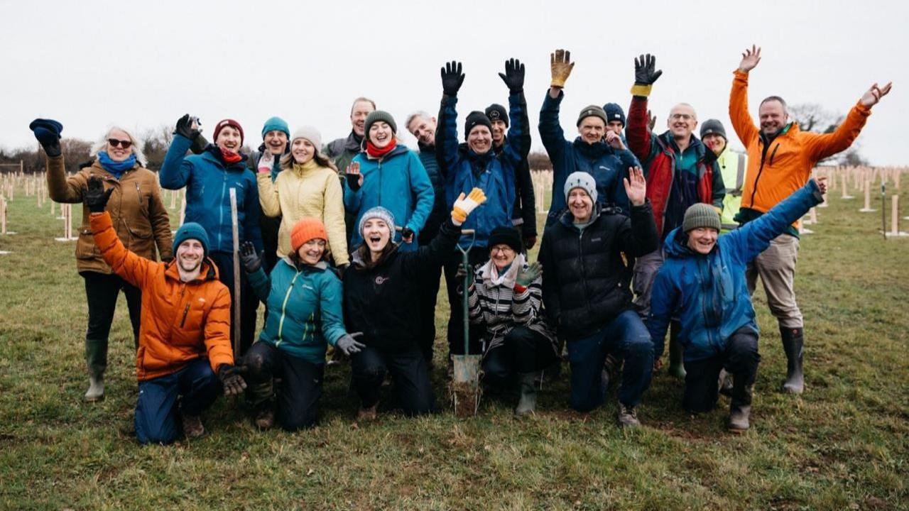 A large group of volunteers wearing rain jackets, gloves and hats. They are standing in a large field surrounded by tree saplings which have just been planted, and are being held up with plastic tree guards. It is a misty, wet day but the group are smiling and waving their arms in the air.