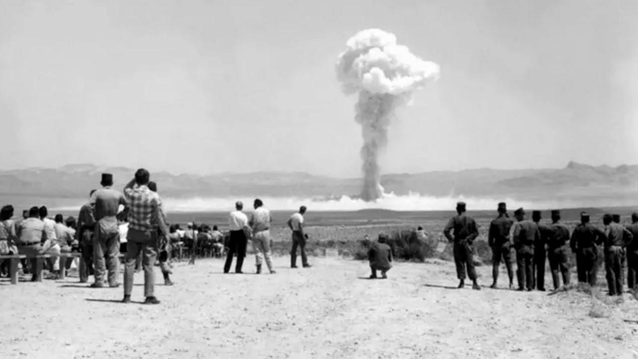 A mushroom cloud erupts in the distance as dozens of people, wearing no protective clothing, stand and watch on a dusty plain.