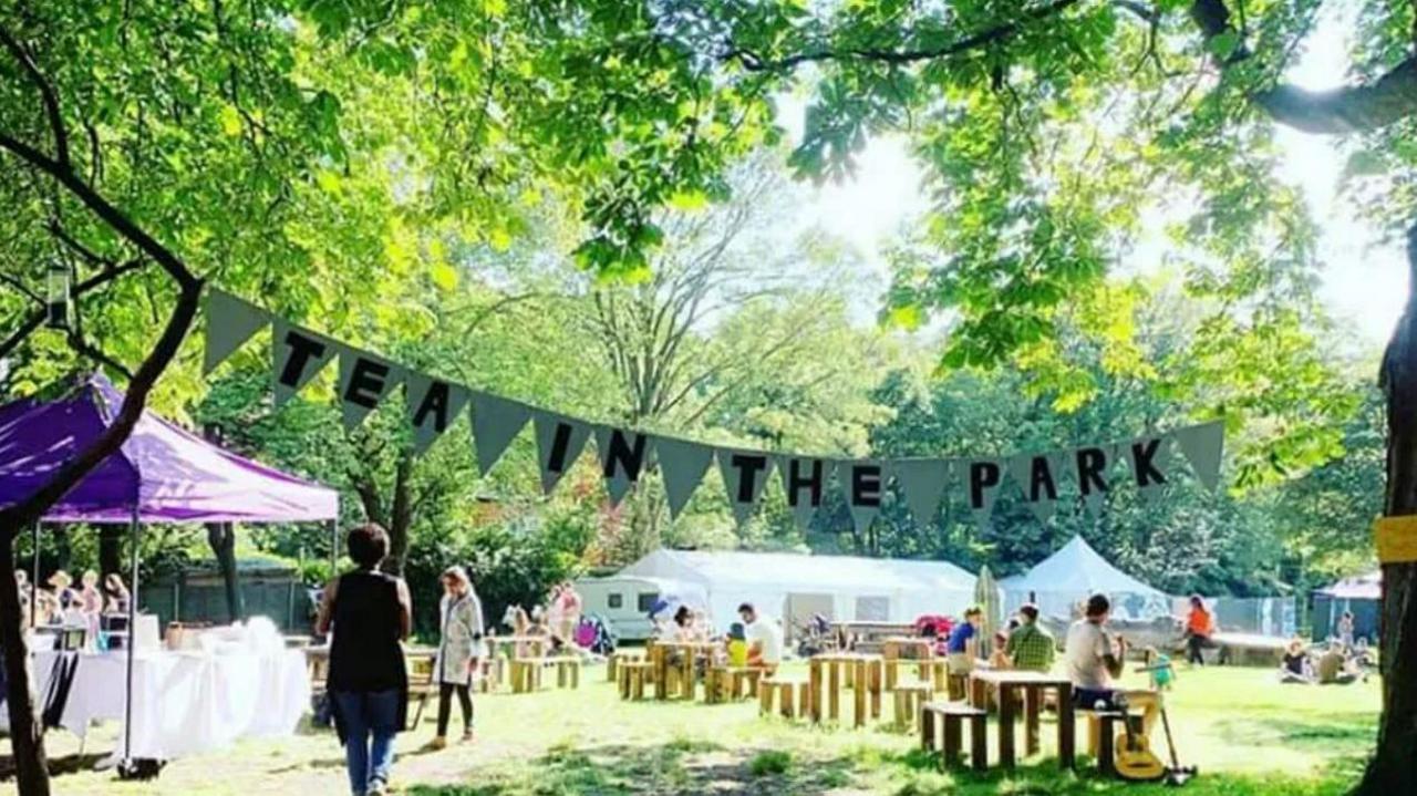 A park on a sunny day. There are white stalls and picnic tables on the grass, with people walking around and sitting at the tables. There is bunting hung from the trees that says "TEA IN THE PARK" in capital letters.