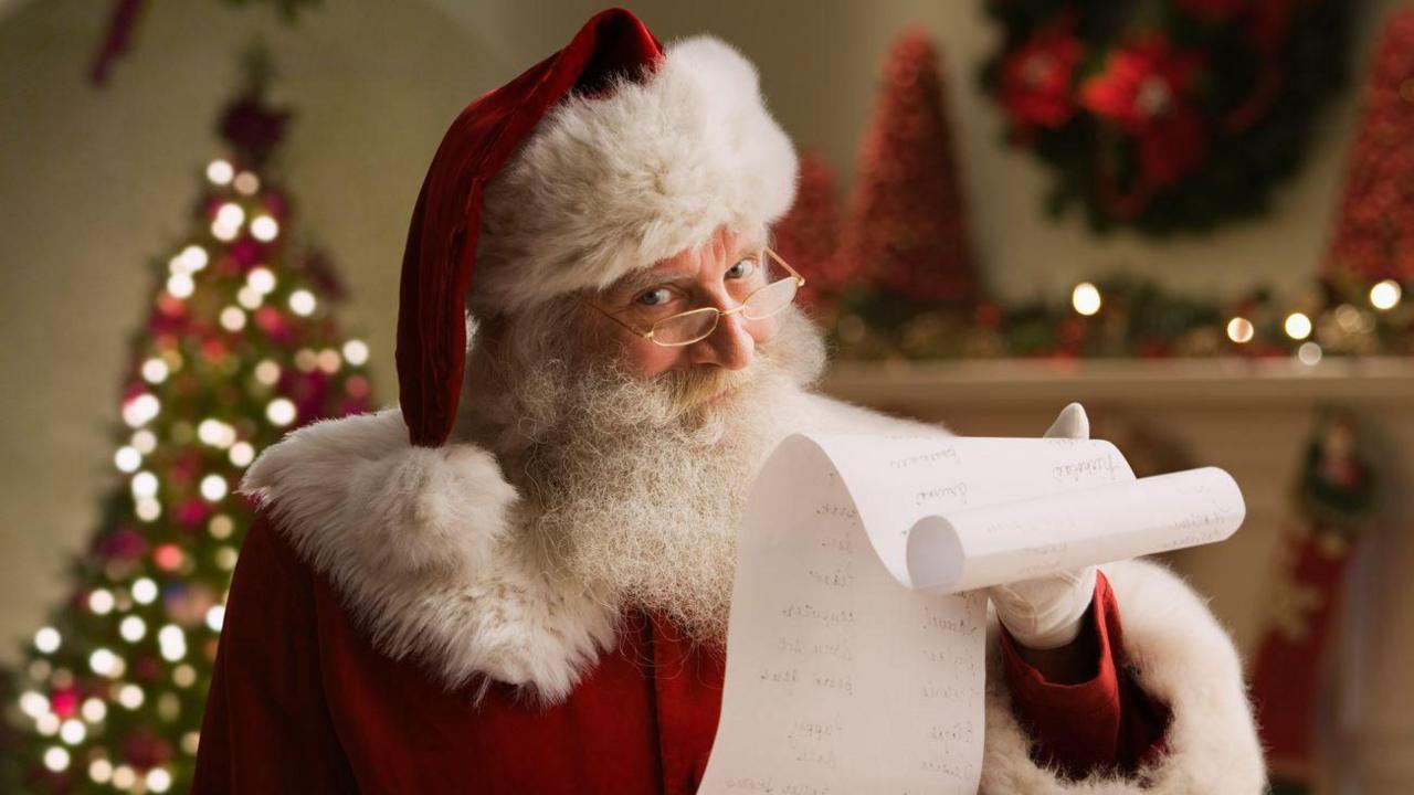 A man with a white beard and glasses wearing a red cloak and red hat with white fur trim, holding a long roll of paper with Christmas lights behind him
