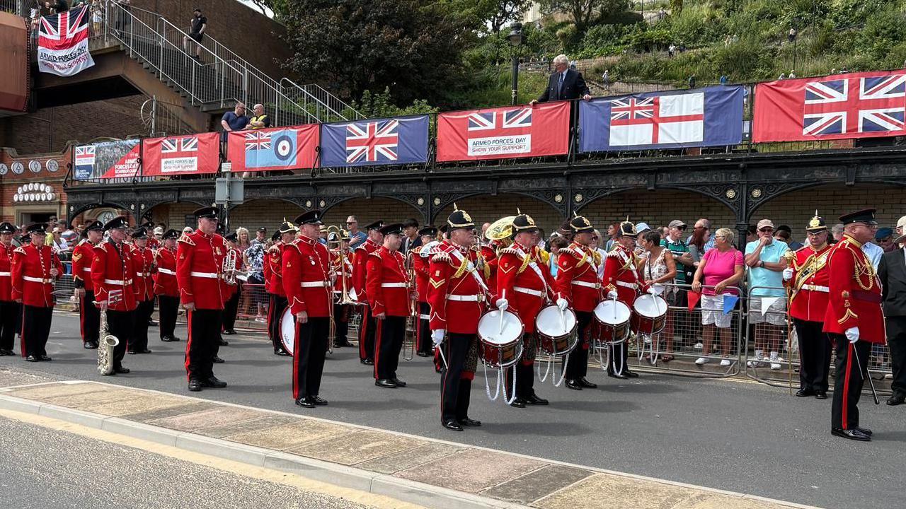 A large military band parading: a large group of men in red uniforms, four at the front with drums.