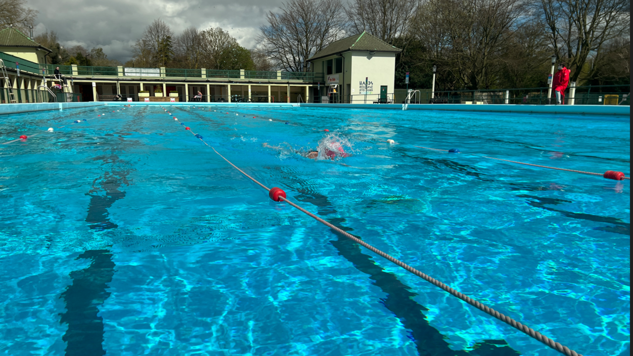 An outdoor lido swimming pool. At least one person is swimming lengths. There are ropes dividing up the lanes.