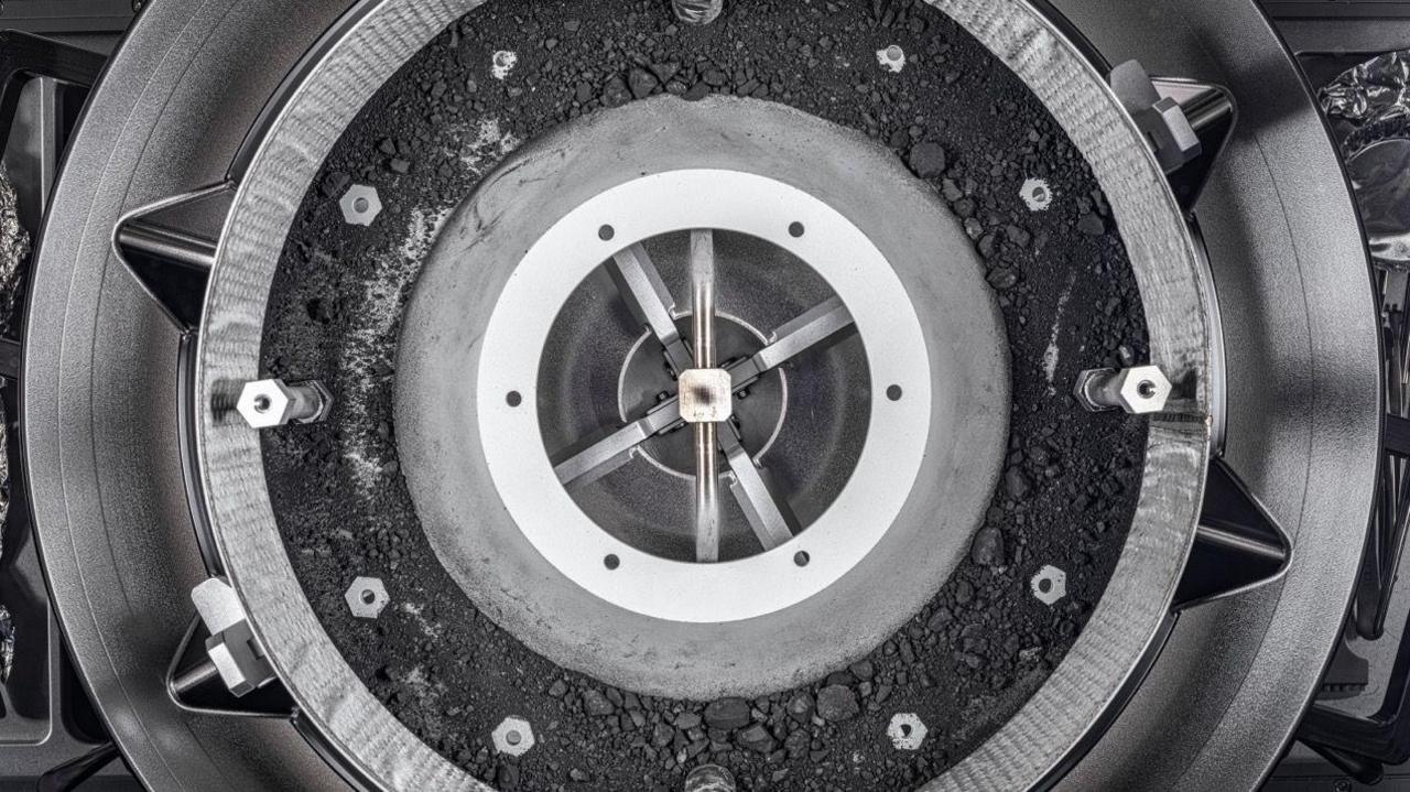 Nasa capsule that looks like an alloy wheel containing the black dusty sample of asteroid Bennu in a central section. The sample resembles coal dust with different sizes of fragments of black rock. There are also a number of bolts that were holding the capsule's lid in place. 