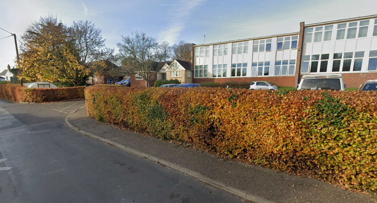 A 1970s-style school building with a bank of white-framed windows and flat roof, viewed from a roadside with head-height hedging in between.