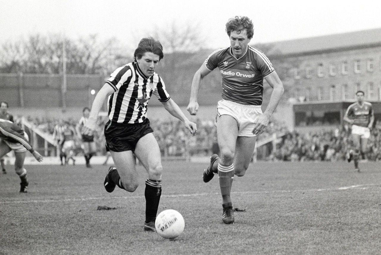 A black and white picture of Terry Butcher, of Ipswich Town, chasing alongside Peter Beardsley of Newcastle United, who has the ball at his feet. Open terracing at St James' Park is in the background, with flats behind the ground.
