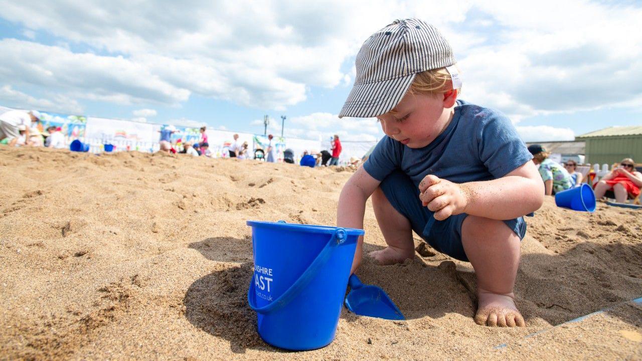 A toddler wearing a blue top and stripy hat is putting a blue plastic spade into the sand next to a blue bucket. Several people are sat relaxing in the distance on the beach.