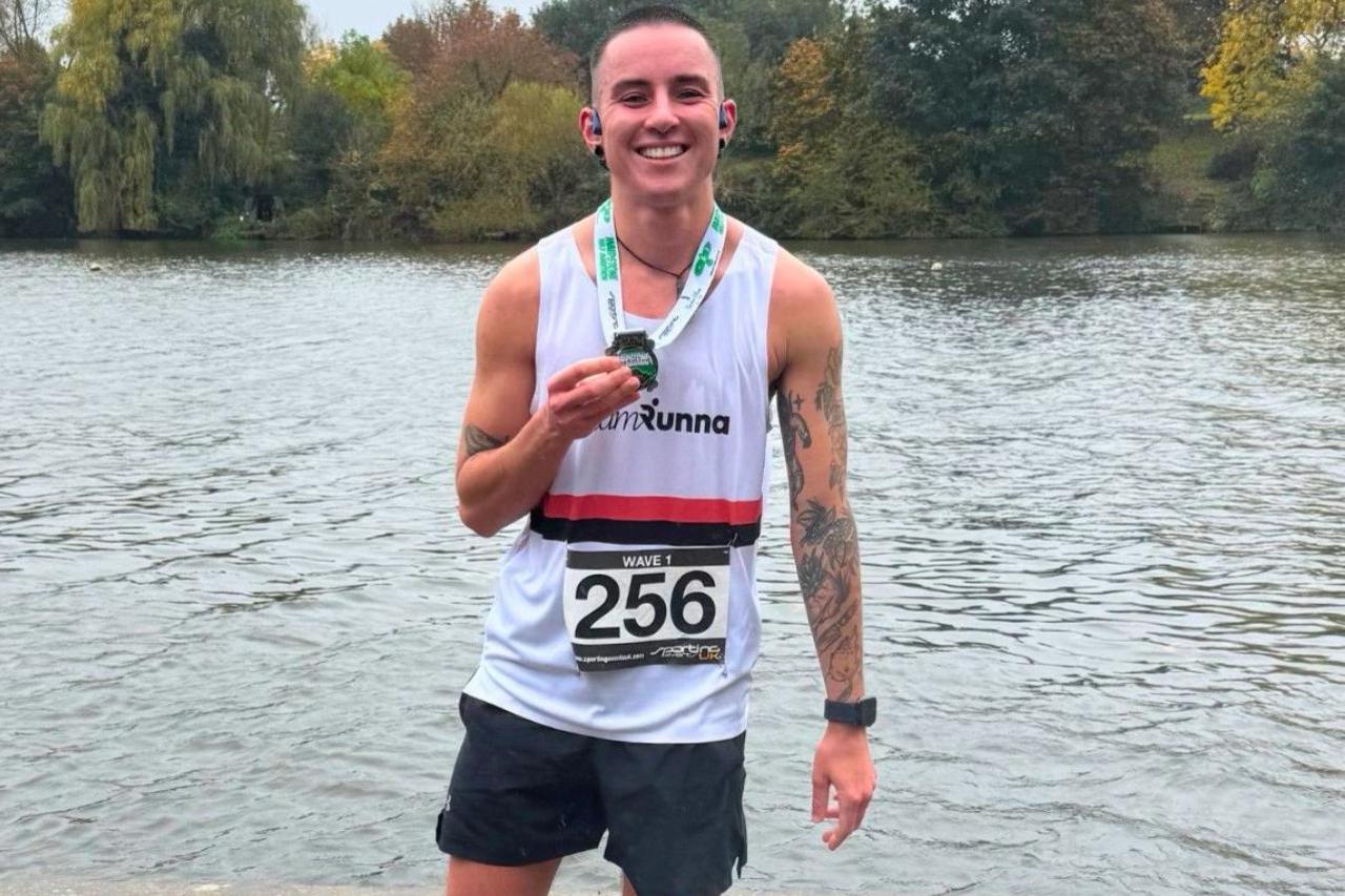 A man is standing in front of a lake. He is wearing running clothes and a bib with "256 on" and is smiling at the camera. He is holding up a medal